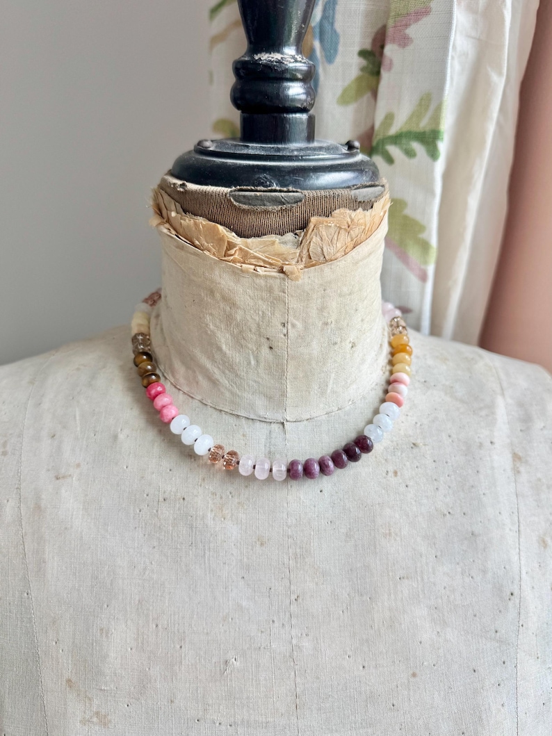 Op de afbeelding: Een kralenketting met een verscheidenheid aan gladde, ronde edelsteenkralen in tinten roze, wit, dieppaars, bruin en doorschijnend amber. De kralen zijn gerangschikt in een doorlopend cirkelvormig patroon rond de nek van een vintage paspop.