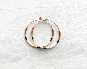 Könnte beinhalten: Goldene Creolen-Ohrringe mit einem Perlenmuster in Creme, Schwarz, Braun und Gold. Die Ohrringe werden auf einer weißen Holzoberfläche präsentiert. Modischer Ohrschmuck.