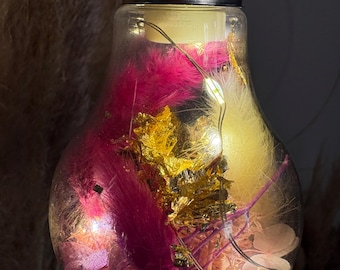 May include: A clear glass lightbulb filled with pink and yellow feathers, gold leaf, and tiny white lights. The bulb has a black base and is lit from within, creating a warm glow. Decorative lighting.