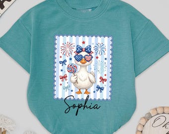 May include: A teal baby romper featuring a graphic of a cartoon duck wearing sunglasses and a bow, holding a lollipop. The graphic has a patriotic theme with fireworks and bows. The name "Sophia" is written below the graphic.