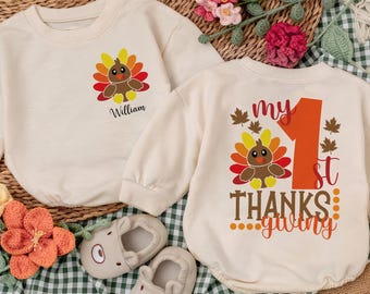 May include: Two off-white baby outfits with Thanksgiving-themed turkey graphics. One says "William" and the other says "my 1st Thanksgiving". The outfits are paired with baby shoes and a wooden toy.