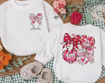 May include: Two white baby onesies with Valentine's Day designs. One features a pink bow and the name "Amelia." The other has hearts, bows, and the phrases "XOXO" and "Be Mine." The image includes baby shoes and floral accents.