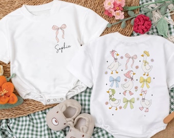 May include: Two white baby onesies with short sleeves. One has a pink bow and the name "Sophia". The other has a pattern of geese with bows. Beige baby shoes and a wooden toy are also in the image. The background is a mix of woven and checkered fabrics.