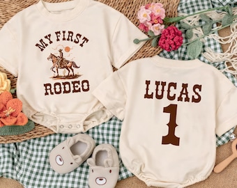 May include: Two cream-colored short-sleeve rompers. One features a brown graphic of a cowboy on a horse with the text "MY FIRST RODEO". The other features the name "LUCAS" and the number "1" in brown text. Includes a pair of beige bear-faced shoes.