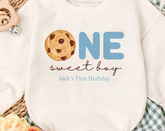 May include: Cream-colored baby romper with the text "ONE sweet boy" and a cookie graphic. The romper is part of a set that includes matching baby shoes. The text "Jack's First Birthday" is also printed on the romper.