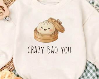 May include: Cream-colored baby onesie with a graphic of a smiling bao bun in a bamboo steamer. The text "CRAZY BAO YOU" is printed below the graphic. The onesie is paired with matching baby shoes.