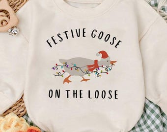 May include: Cream-colored baby romper with long sleeves. The front features a graphic of a goose wearing a Santa hat and wrapped in Christmas lights, with the text "FESTIVE GOOSE ON THE LOOSE". Includes matching baby slippers.