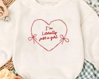 May include: Cream-colored baby romper with long sleeves. A red heart with the text "I'm Literally just a girl" is printed on the front. The romper is paired with beige baby shoes and a wooden toy.