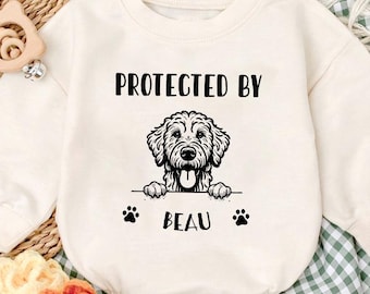 May include: Cream-colored baby onesie with the text "PROTECTED BY" above a black and white dog illustration. Below the dog is the name "BEAU" and paw prints. The onesie is on a green and white checkered blanket.