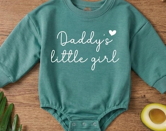 May include: A teal long-sleeve baby bodysuit featuring a ribbed crew neckline and cuffs. The front displays the white text "Daddy's little girl" with a small heart icon above the word "Daddy's". The garment includes three metal snaps at the crotch.