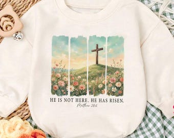May include: Cream-colored baby romper with a watercolor design of a cross on a hill, surrounded by flowers. The text "HE IS NOT HERE. HE HAS RISEN. Matthew 28:6" is printed below the image. The romper is paired with matching baby shoes.