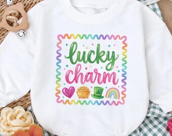 May include: White baby onesie with long sleeves. The front features a colorful graphic that says "lucky charm" with a heart, pot of gold, leprechaun hat, and rainbow. The onesie is surrounded by decorative elements, including flowers and baby shoes.