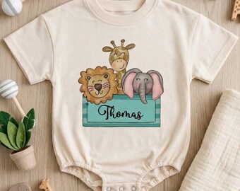 May include: A cream-colored short-sleeve baby bodysuit featuring a graphic illustration of a lion, giraffe, and elephant inside a teal box with the name Thomas written in black script. The garment has a round neckline and snap closures.