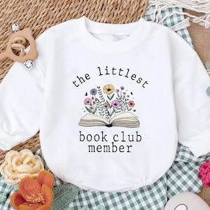 May include: A white long-sleeved sweatshirt featuring a graphic of an open book with colorful wildflowers growing out of the pages. The text above and below the illustration reads, "the littlest book club member" in a simple black font.