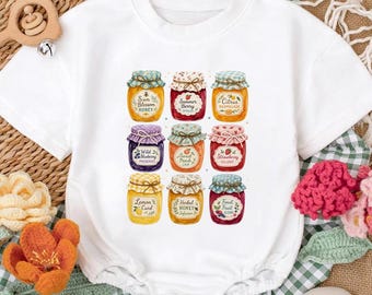 May include: White short-sleeved baby bodysuit featuring a colorful graphic of nine jars of preserves. Each jar is labeled with a different flavor, such as "Summer Berry" and "Strawberry". The jars have decorative lids and labels.