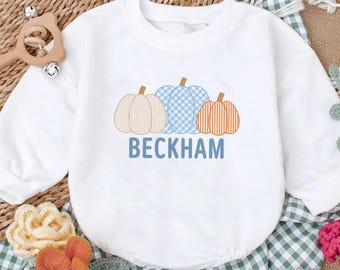May include: A white long-sleeved sweatshirt featuring a graphic of three pumpkins in beige, blue, and orange with striped and checkered patterns. The name BECKHAM is printed in blue capital letters underneath the pumpkins on the front of the garment.