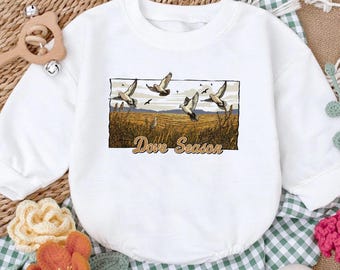 May include: A white long-sleeve sweatshirt featuring a rectangular graphic print of birds flying over a golden field with the text Dove Season written in a brown script font below the illustration. The garment has a crew neckline and ribbed cuffs.
