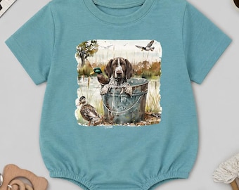 May include: A teal baby romper with a watercolor illustration of a dog in a bucket, surrounded by ducks and birds. The word "BABY" is spelled out in wooden letters above the romper. Includes a wooden toy, sunglasses, and a rainbow.