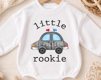 May include: A white long-sleeve baby romper featuring a graphic of a grey police car with blue windows and a red and blue light bar on top. The text "little" is printed in black above the car, and the word "rookie" is printed in black below it.