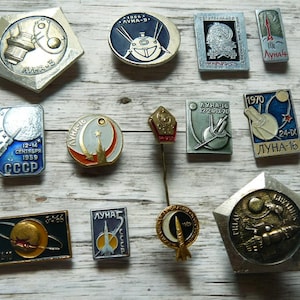 Könnte beinhalten: Eine Sammlung von Vintage-Anstecknadeln und -Abzeichen zum Thema Weltraum. Designs zeigen Raketen, Planeten und den Mond, mit kyrillischem Text. Die Anstecknadeln sind in verschiedenen Formen und Größen gehalten, mit Silber-, Gold- und Blautönen.