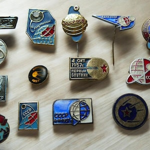 Può includere: Una collezione di spille e distintivi vintage dell'era sovietica, con vari disegni relativi all'esplorazione spaziale e all'URSS. Le spille sono di varie forme, tra cui rotonde, quadrate e rettangolari, con finiture in smalto e metallo. Alcune spille hanno testo.