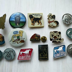 Puede incluir: Una colección de pines y chapas vintage con temática de perros. Los pines presentan varias razas de perros, incluyendo un Mastín, un Bloodhound y un Dachshund. Algunos pines tienen texto, incluyendo "CANIS" y "RSCA".