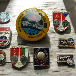 Könnte beinhalten: Eine Sammlung von Vintage-Anstecknadeln und -Abzeichen zum Thema Weltraum, mit verschiedenen Designs mit Text und Bildern, die sich auf die Apollo-Sojus-Mission und die sowjetischen Weltraumprogramme beziehen. Die Anstecknadeln sind in verschiedenen Formen und Farben.