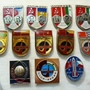 May include: A collection of vintage Soviet-era pins and badges. The pins feature various designs, including portraits, flags, and space-themed imagery. The color palette includes red, gold, blue, and white. Some pins have the text "CCCP" and "INTERKOSMOS".