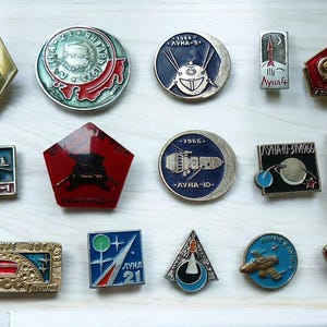 Pode incluir: Uma coleção de pins vintage com tema espacial em várias formas. Os pins apresentam foguetes, planetas e texto cirílico, com cores como ouro, vermelho, azul e prata. Alguns têm o texto "LUNA".