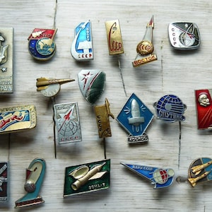 Pode incluir: Uma coleção de pins vintage da era soviética com tema espacial. Os pins apresentam foguetes, satélites e cosmonautas em várias formas e cores, incluindo ouro, azul, vermelho e prata. Alguns pins têm texto cirílico.