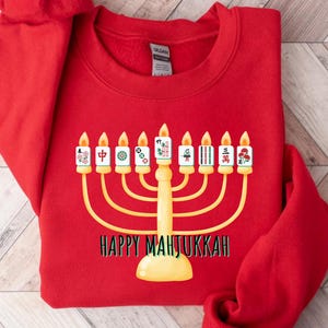 May include: A red sweatshirt featuring a gold menorah design with lit candles and Mahjong tiles. The text "HAPPY MAHJUKKAH" is printed below the menorah in black. The sweatshirt is made of a soft material.