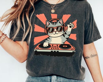 Puede incluir: Camiseta gris oscuro con un gato de dibujos animados DJ con gafas de sol y auriculares, saludando con la pata. El gato está frente a un diseño de rayos de sol rojo y una mesa de mezclas de DJ. Prenda casual.