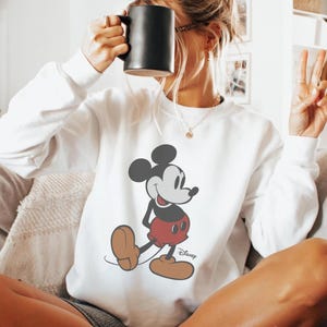 Puede incluir: Sudadera blanca con un dise&ntilde;o de Mickey Mouse. El personaje de dibujos animados est&aacute; representado en negro, rojo y marr&oacute;n. El logotipo de Disney est&aacute; impreso debajo. La sudadera tiene mangas largas y cuello redondo.