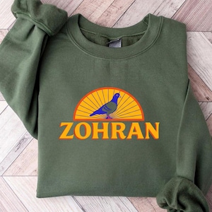 May include: An olive green sweatshirt featuring a graphic of a blue pigeon perched on a yellow and orange sunburst design. The name "ZOHRAN" is printed in orange below the graphic. The sweatshirt is made of a soft material.