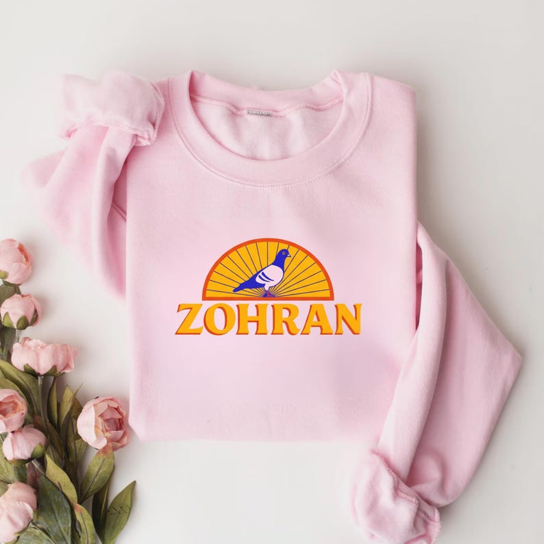 May include: A light pink sweatshirt featuring a graphic of a blue pigeon inside a yellow and orange semi-circle, with the word "ZOHRAN" in orange block letters below. The sweatshirt has long sleeves and a crew neck.