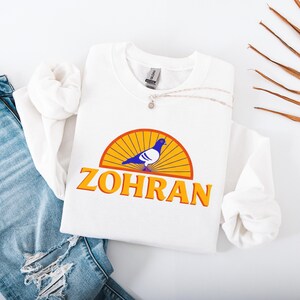 May include: White crewneck sweatshirt with a graphic featuring a blue and white pigeon inside a yellow and orange semi-circle, with the word "ZOHRAN" in orange. A silver necklace with a small pendant rests on the sweatshirt.