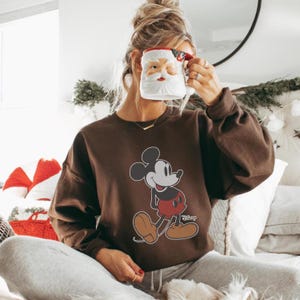 Puede incluir: Una sudadera marr&oacute;n con un gr&aacute;fico de Mickey Mouse. El logotipo de Disney es visible. La persona sostiene una taza de Pap&aacute; Noel. La sudadera es de corte holgado. La imagen est&aacute; tomada en un ambiente relajado.
