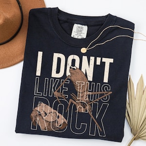 May include: Navy blue t-shirt with the text "I DON'T LIKE THIS ROCK" and a graphic of a bug and a rock. A gold necklace and a brown hat are also in the image.