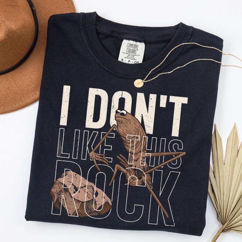 May include: Navy blue t-shirt with the text "I DON'T LIKE THIS ROCK" and a graphic of a bug and a rock. A gold necklace and a brown hat are also in the image.