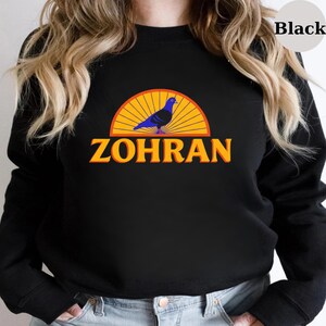 May include: Black sweatshirt featuring a graphic design. The design includes a blue pigeon, a yellow and orange sunburst, and the word "ZOHRAN" in orange letters. The sweatshirt is a solid black color.