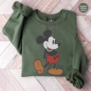 Puede incluir: Sudadera verde militar jaspeada con un gr&aacute;fico cl&aacute;sico de Mickey Mouse. El dise&ntilde;o incluye las orejas negras, la cara, los pantalones cortos rojos y los zapatos marrones de Mickey. El logotipo de Disney es visible. La sudadera tiene cuello redondo y mangas largas.