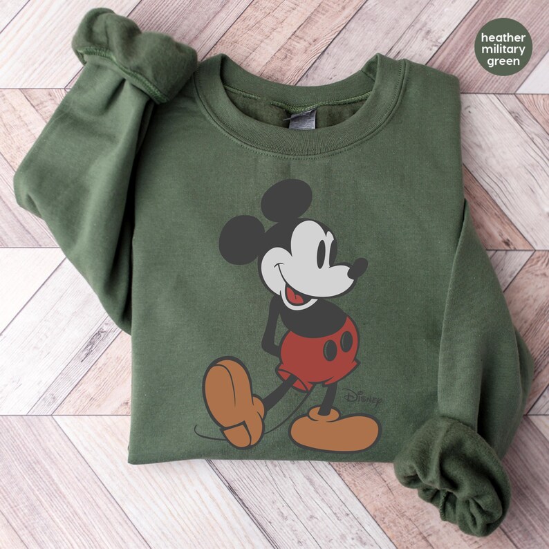 Puede incluir: Sudadera verde militar jaspeada con un gr&aacute;fico cl&aacute;sico de Mickey Mouse. El dise&ntilde;o incluye las orejas negras, la cara, los pantalones cortos rojos y los zapatos marrones de Mickey. El logotipo de Disney es visible. La sudadera tiene cuello redondo y mangas largas.