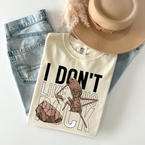 May include: Cream-colored t-shirt with the text "I DON'T LIKE THIS ROCK" and a graphic of an insect and a rock. The shirt is paired with light-wash jeans, a gold necklace, and a tan hat.