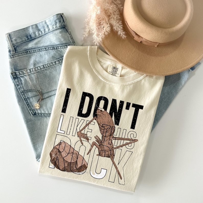 May include: Cream-colored t-shirt with the text "I DON'T LIKE THIS ROCK" and a graphic of an insect and a rock. The shirt is paired with light-wash jeans, a gold necklace, and a tan hat.