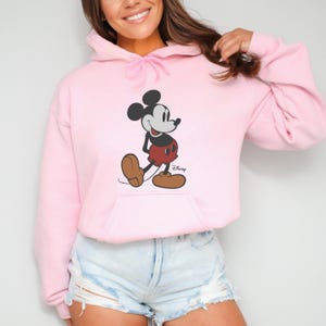 Puede incluir: Sudadera con capucha rosa claro con un dise&ntilde;o de Mickey Mouse. El personaje de dibujos animados est&aacute; en negro, rojo y marr&oacute;n. El logotipo de Disney est&aacute; impreso en la parte inferior derecha. La sudadera tiene una capucha con cord&oacute;n y un bolsillo delantero.