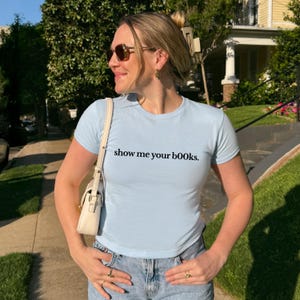 May include: Light blue short-sleeved t-shirt with the text "show me your books." The shirt is paired with blue jeans. A small, cream-colored purse is worn over the shoulder. The person is wearing sunglasses and rings.