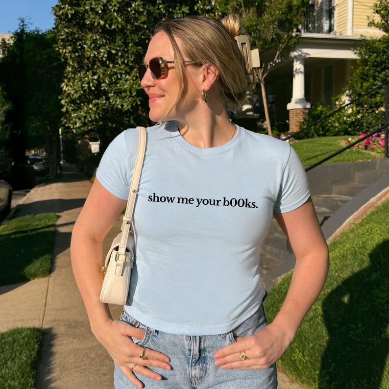 May include: Light blue short-sleeved t-shirt with the text "show me your books." The shirt is paired with blue jeans. A small, cream-colored purse is worn over the shoulder. The person is wearing sunglasses and rings.