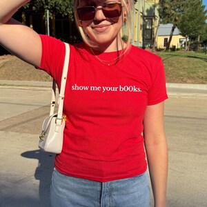 May include: A red t-shirt with the phrase "show me your books" printed in white. The shirt is paired with blue jeans and a white purse. The image is taken outdoors on a sunny day.
