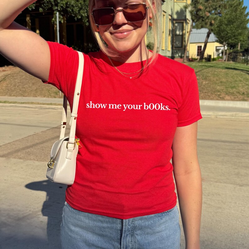 May include: A red t-shirt with the phrase "show me your books" printed in white. The shirt is paired with blue jeans and a white purse. The image is taken outdoors on a sunny day.