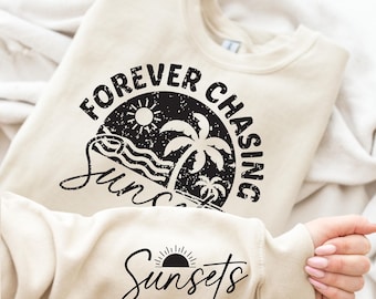 May include: A cream-coloured sweatshirt featuring a black graphic with the words "Forever Chasing Sunsets" encircling a sun, palm trees, and waves. The sleeve displays the word "Sunsets" with a sun graphic. The sweatshirt is folded.